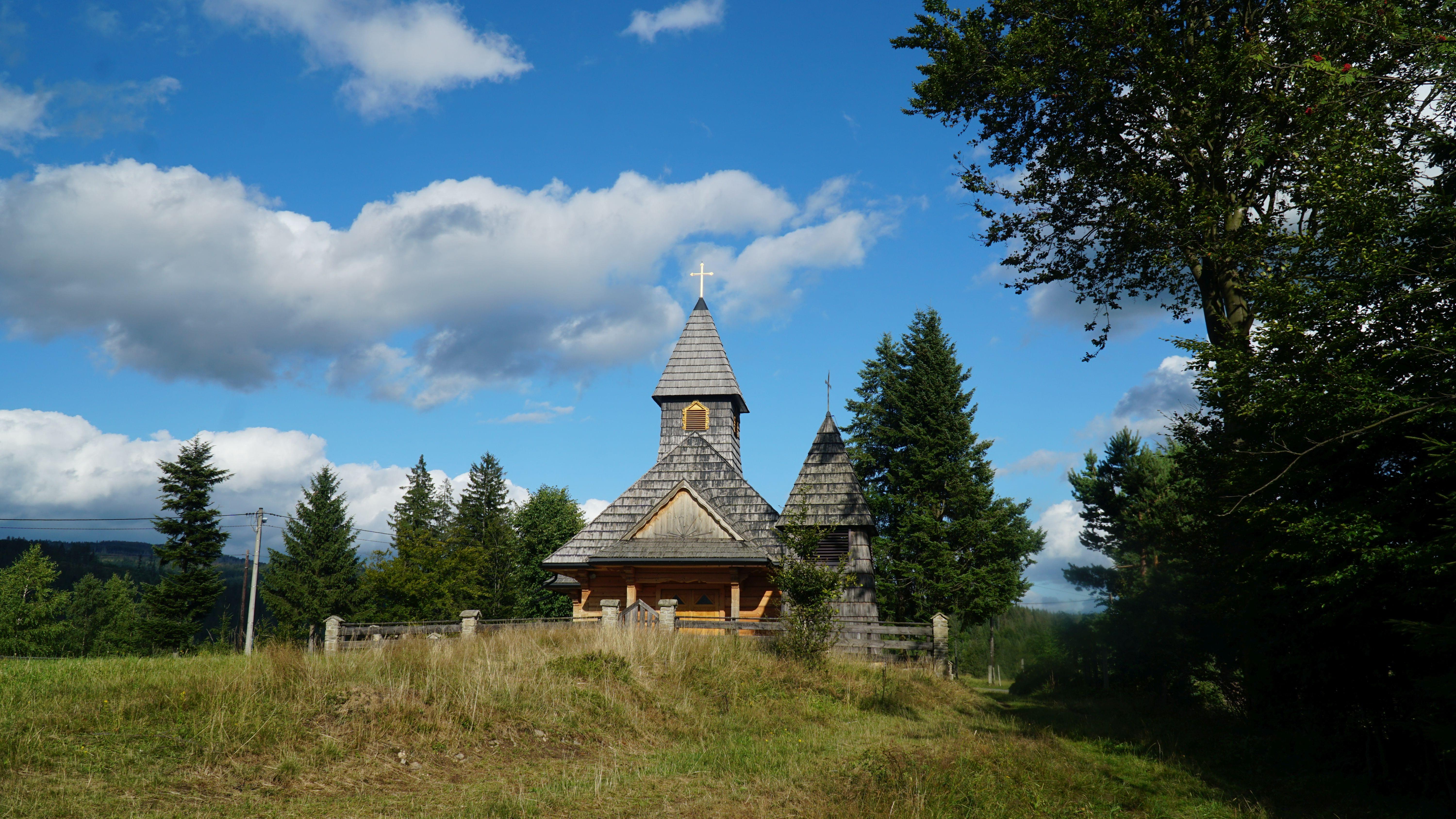Frontowa strona drewnianego kościoła. Dach pokryty gontem. Po prawej stronie utrzymana w
podobnym stylu dzwonnica. Całość okala drewniano-kamienne ogrodzenie.